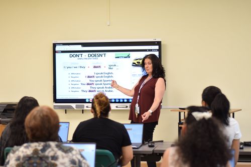 Students in a classroom seen from behind, looking at the blackboard. The teacher is talking to them.