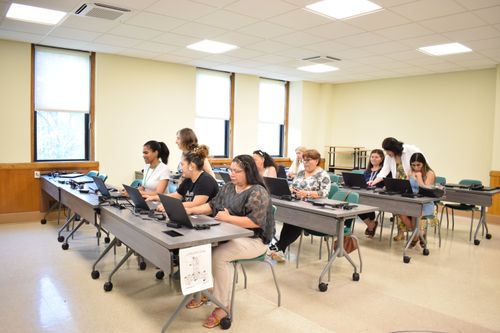 Students are sitting at desks in a large classroom, listening attentively to the teacher.