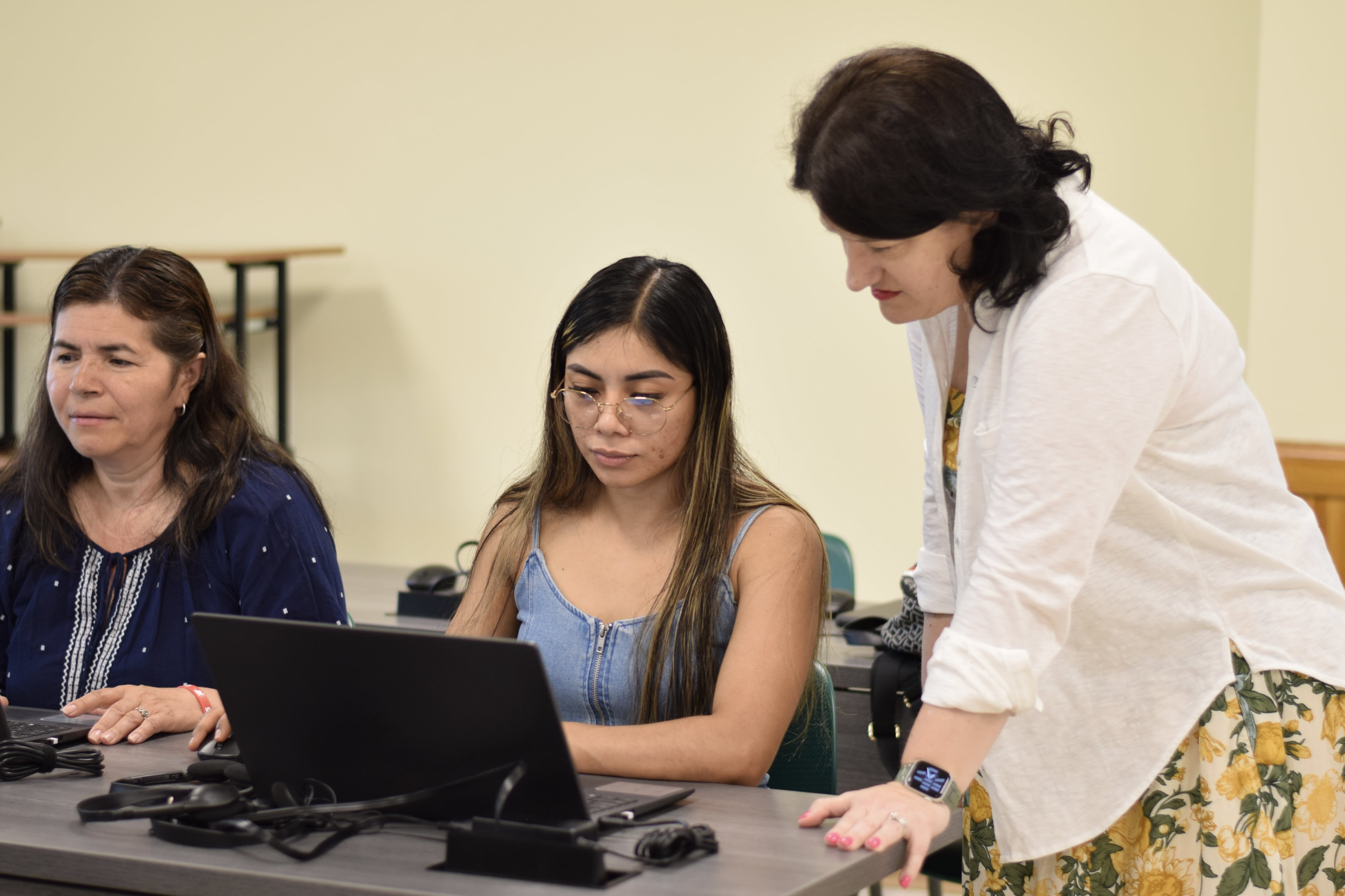 The teacher is assisting a student in a computer classroom, guiding her on the assignment.