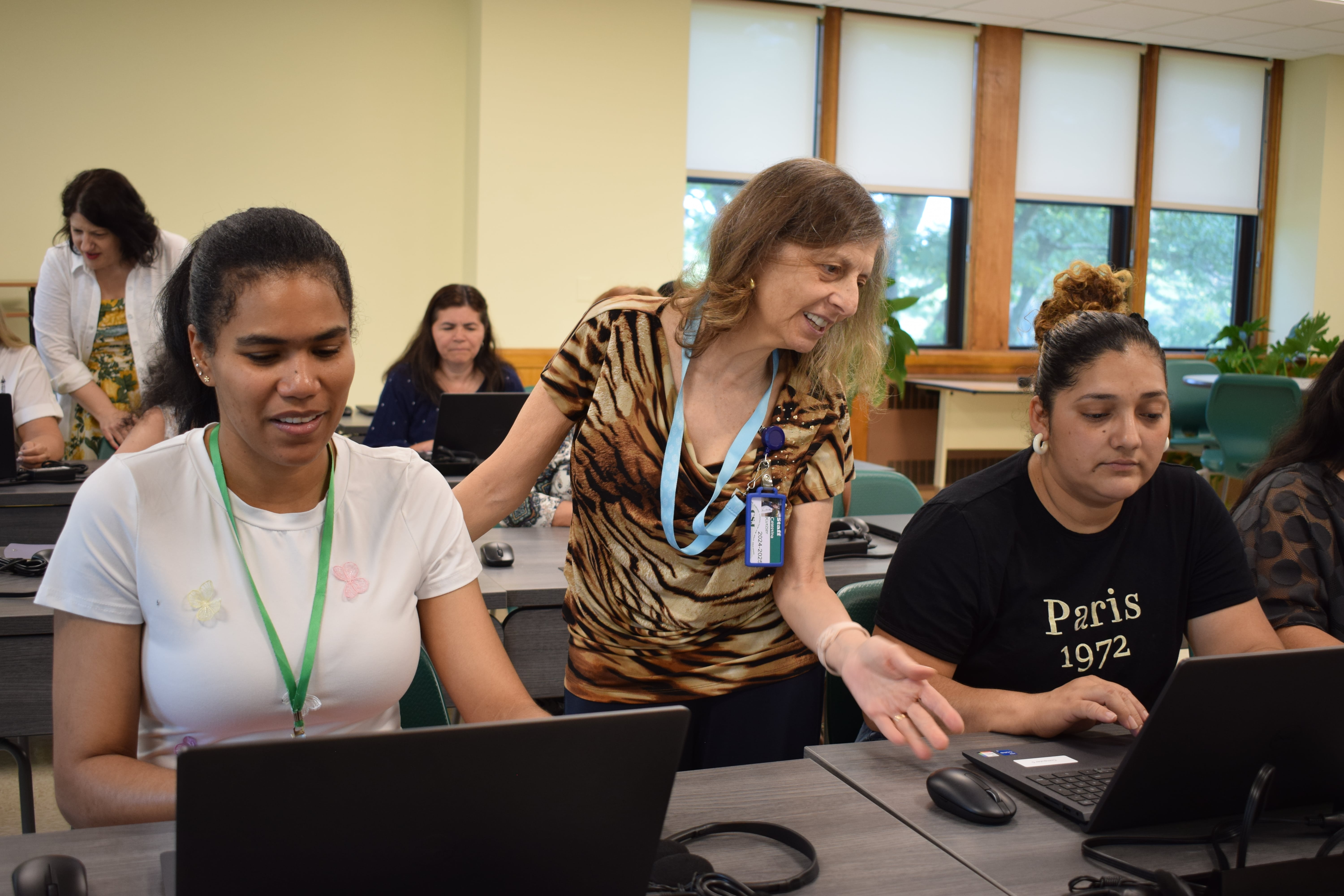 The teacher is assisting two students in a computer classroom, guiding them on their assignments.