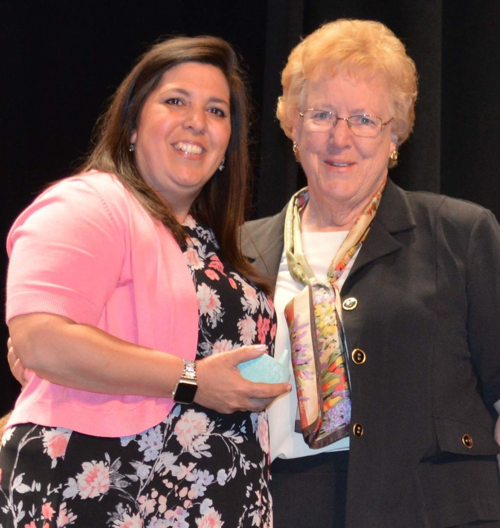 Denise Murray is receiving the torch from Sister Kathleen in a ceremonial handoff, symbolizing the passing of leadership and mentorship.