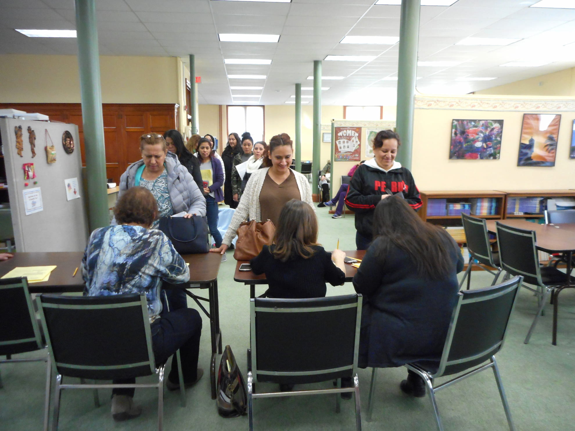 People standing in line at a registration desk inside a spacious room with paintings on the walls and bookshelves.