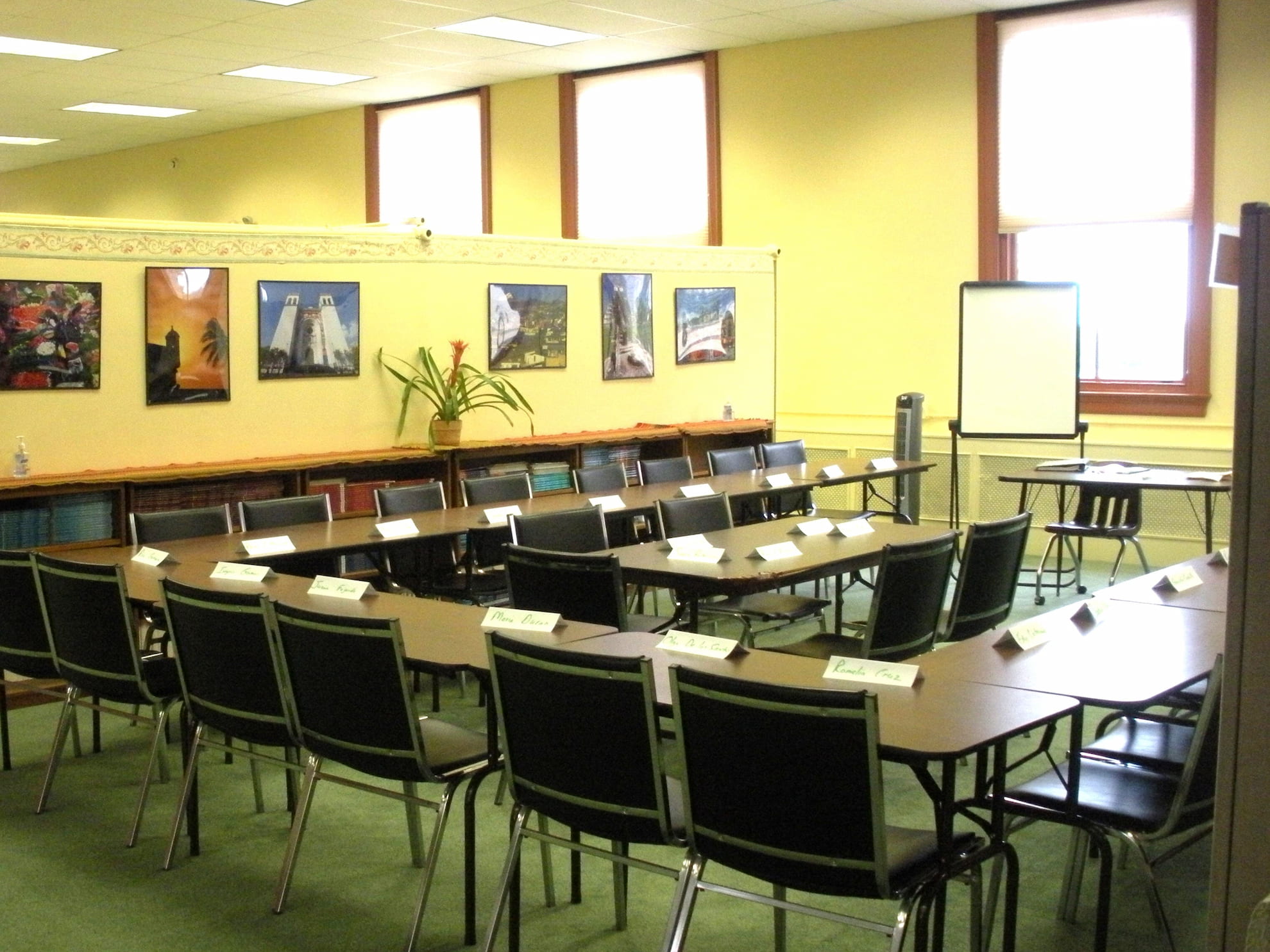 Conference room with U-shaped tables, black chairs, name cards, a flip chart, and framed pictures on yellow walls.
