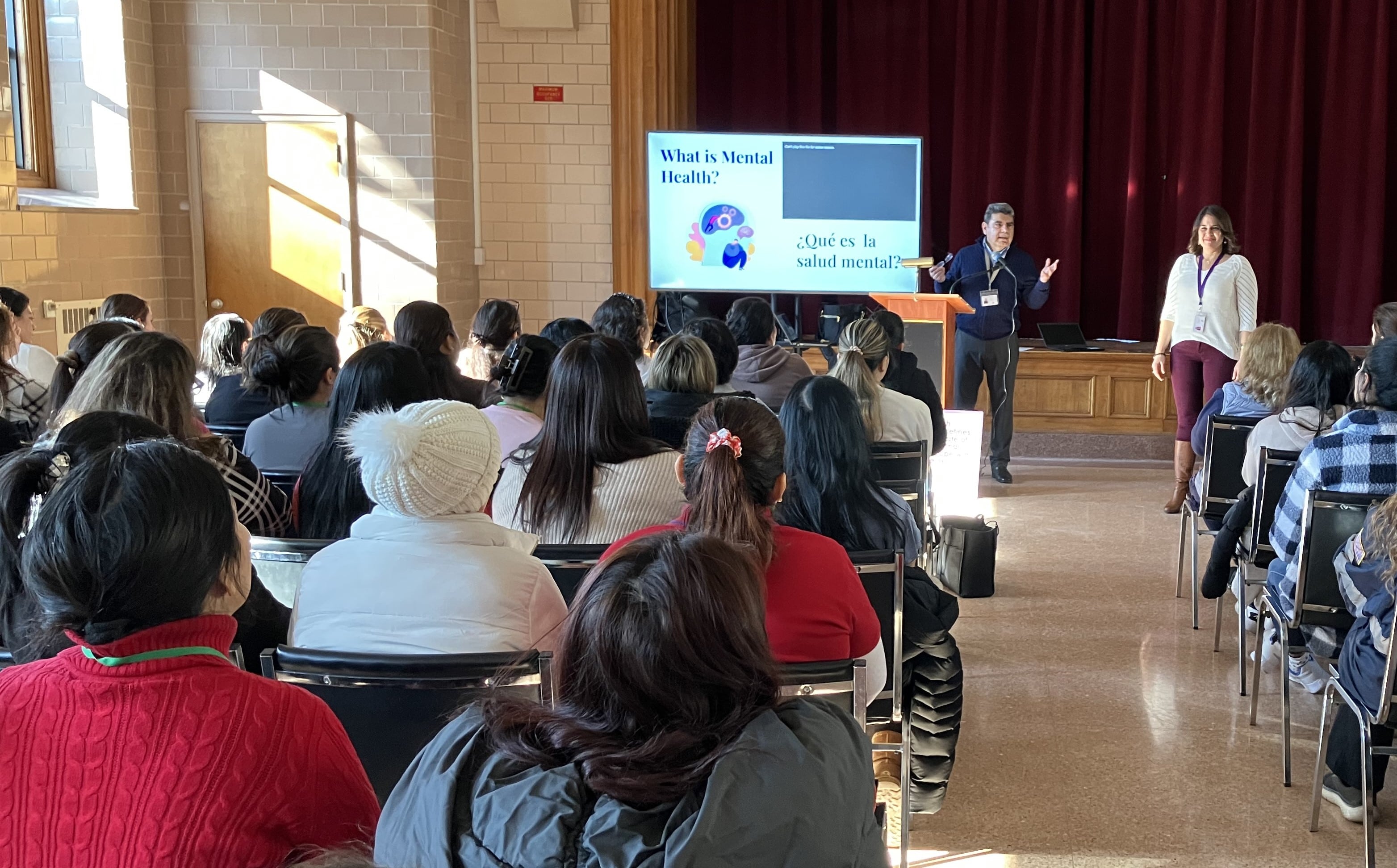 Women listening to a man speaking on stage at a Hispanic counseling center.