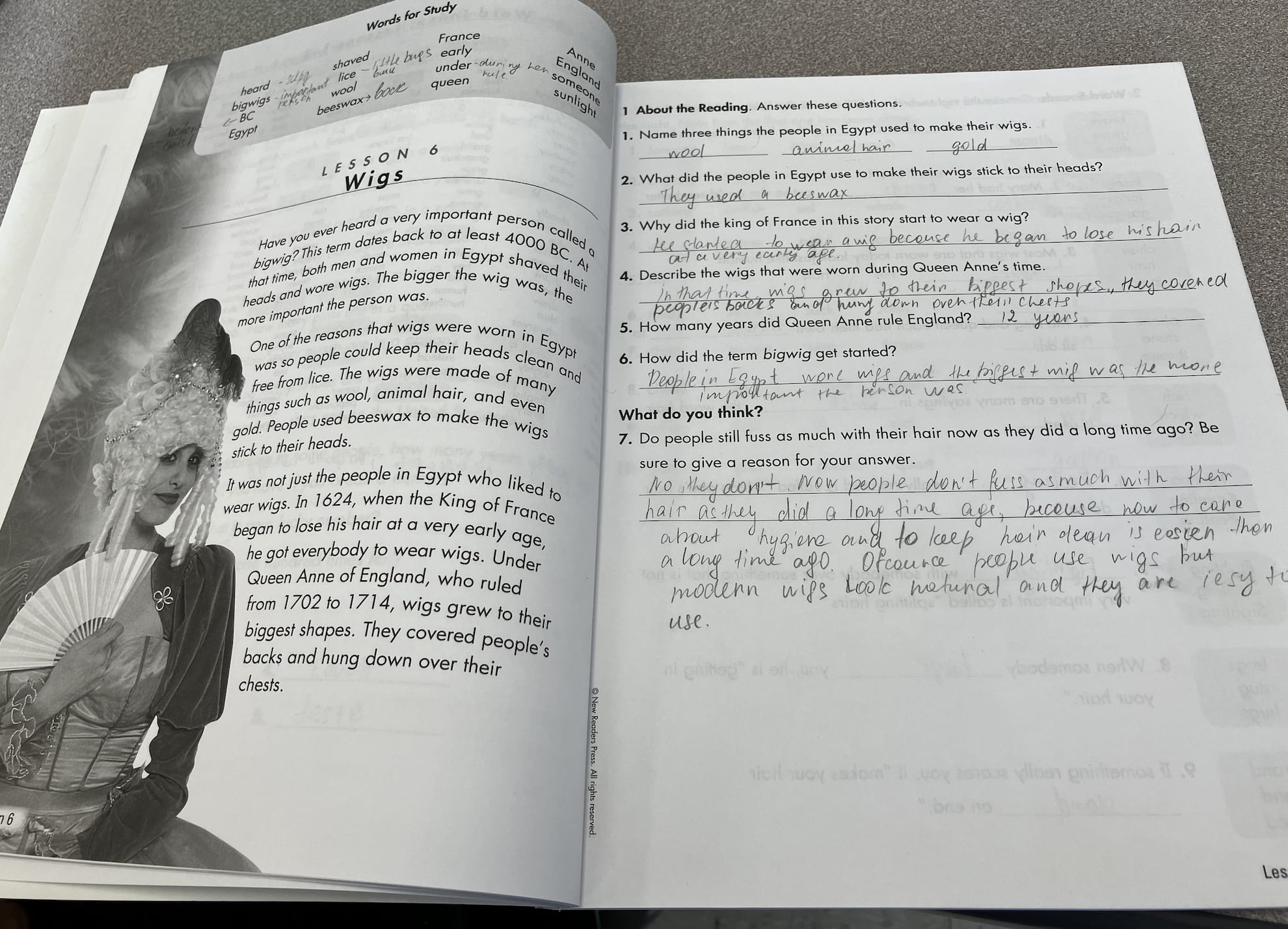 Open textbook showing Lesson 6 titled 'Wigs' with a black and white image of a woman wearing an elaborate historical wig and holding a fan, alongside a questionnaire with handwritten answers about wigs in Egypt and historical figures.
