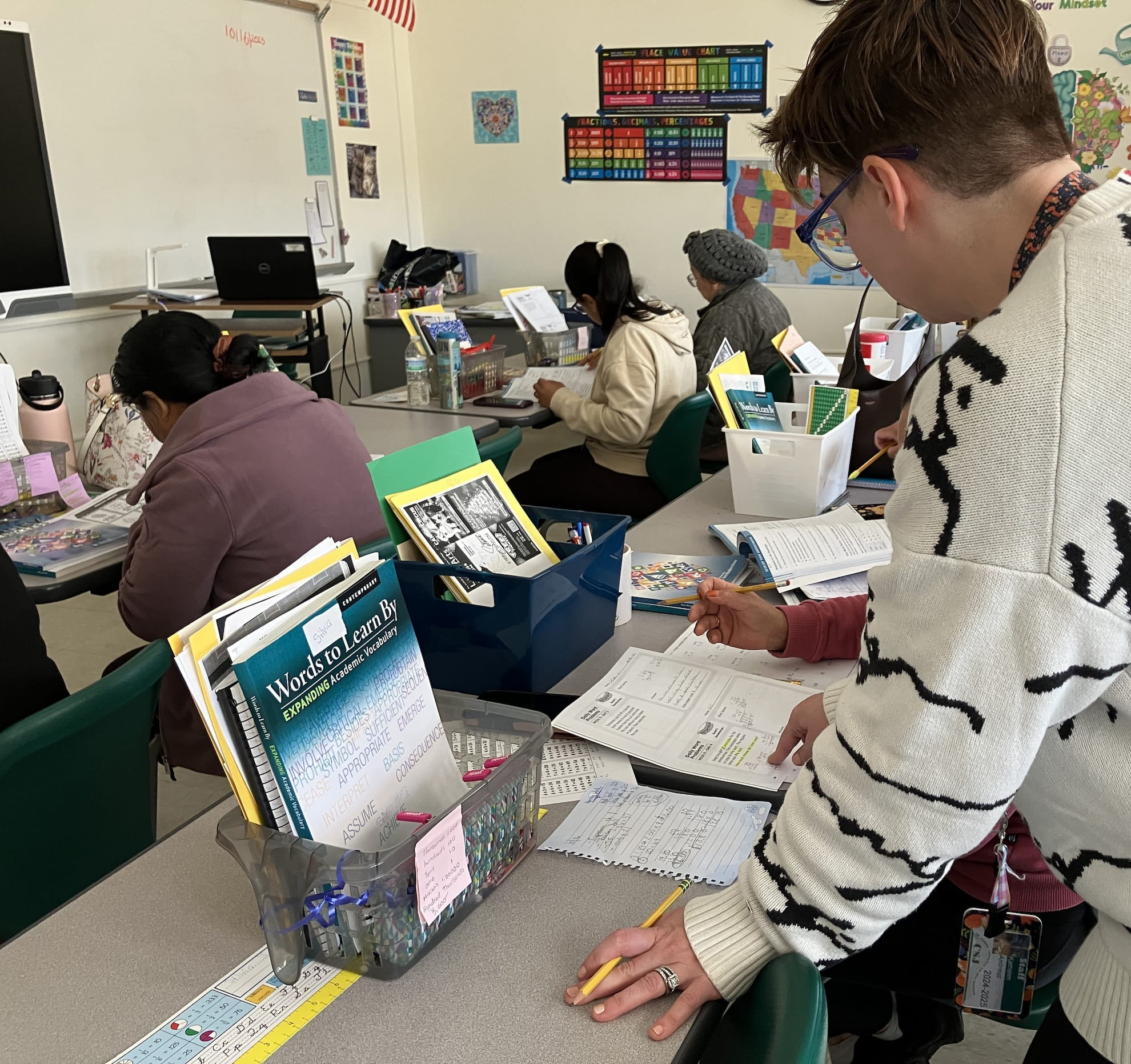 A classroom scene with a teacher assisting a student with worksheets at a desk while other students study in the background.