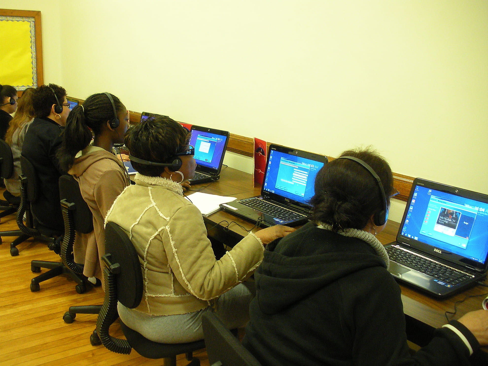 A group of women wearing headsets seated in a row using laptops in a classroom or computer lab.