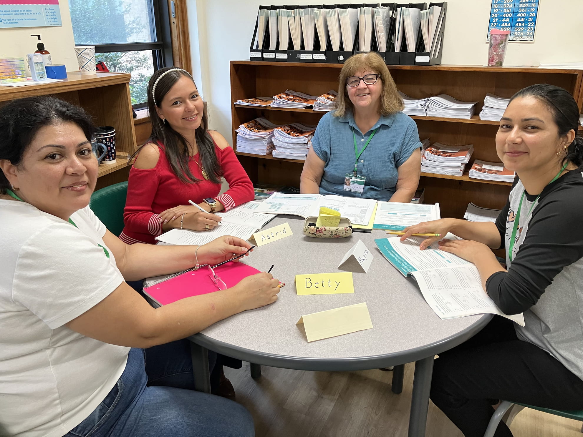 Four women sitting around a table in a study room with open books and name tags, smiling at the camera.