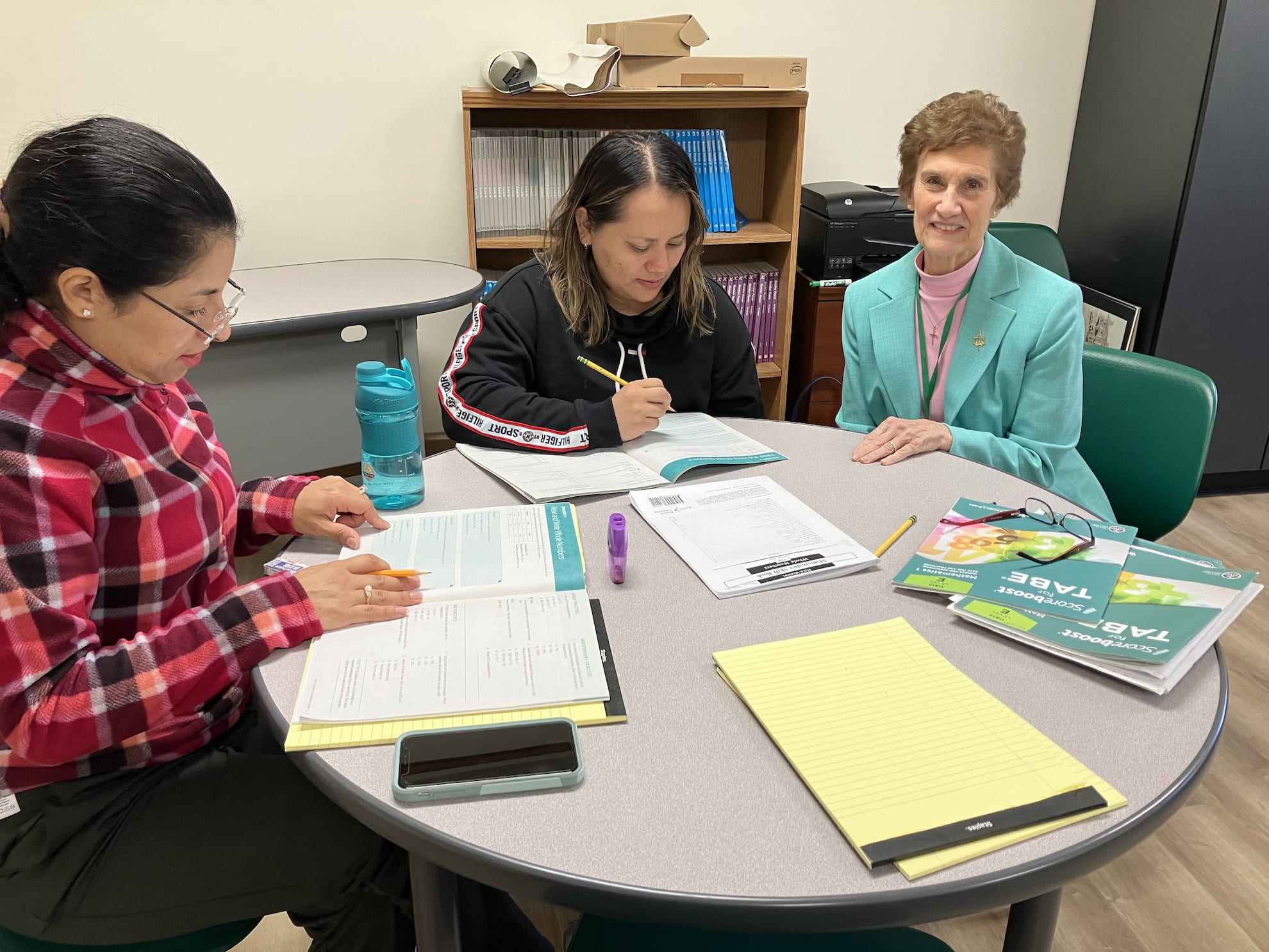 Three women sitting around a round table with papers, notebooks, and pens, engaged in a discussion or study session in an office setting.