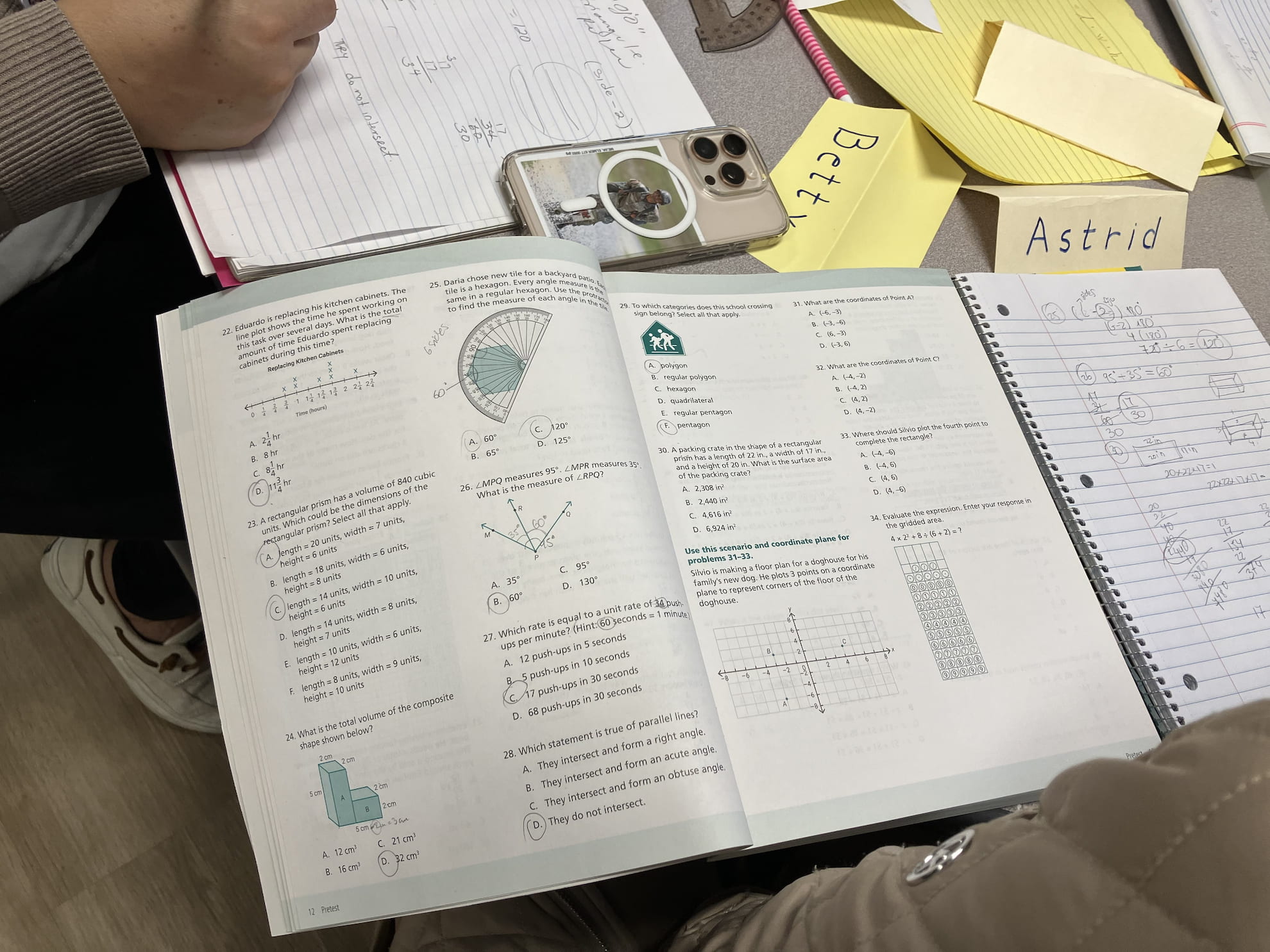 Open math workbook with geometry and volume problems, a notebook with handwritten calculations, a smartphone, and name tags labeled Betty and Astrid on a desk.