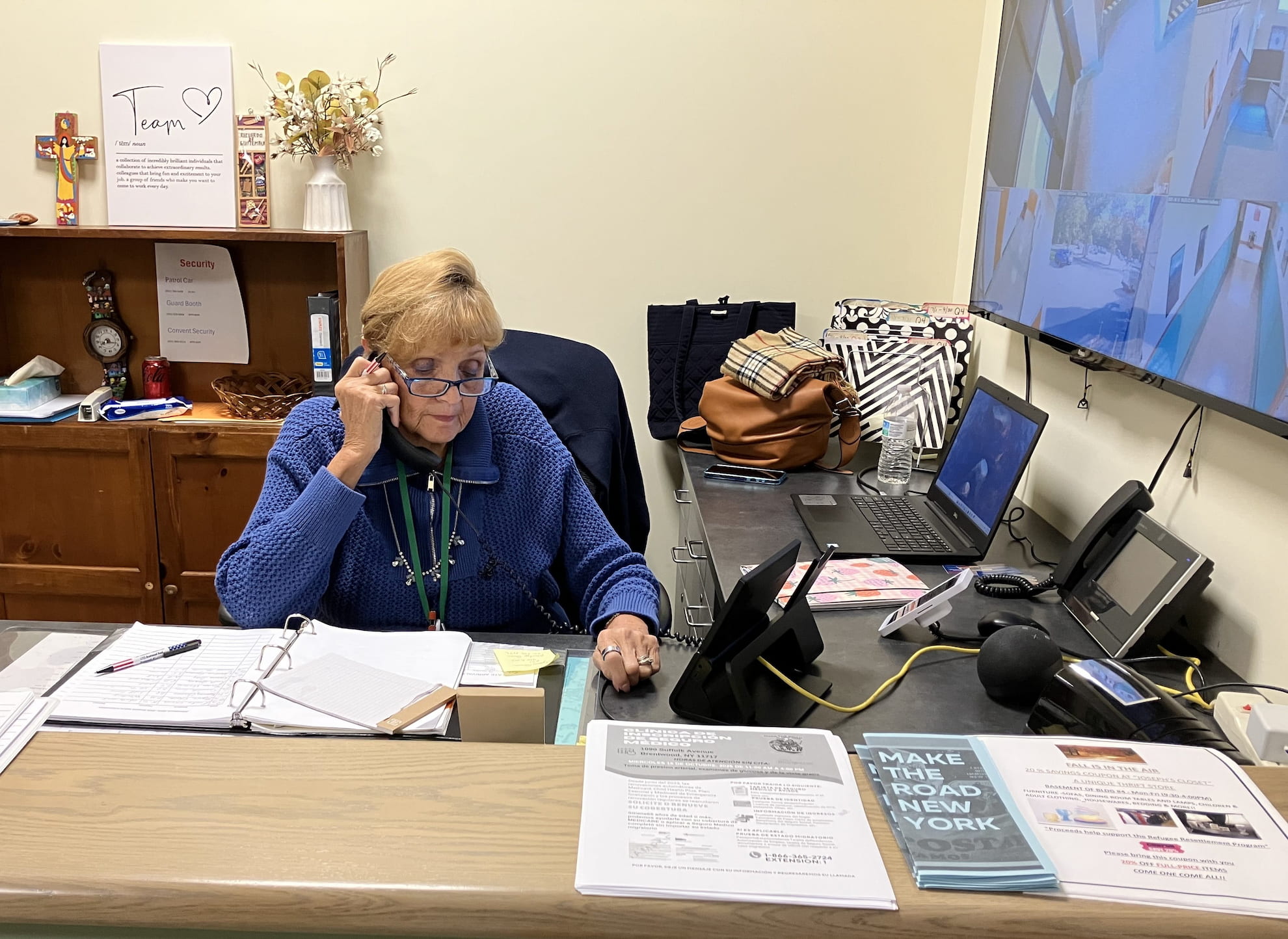 Older woman in blue sweater speaking on the phone at an office desk with paperwork, a laptop, and security monitors.