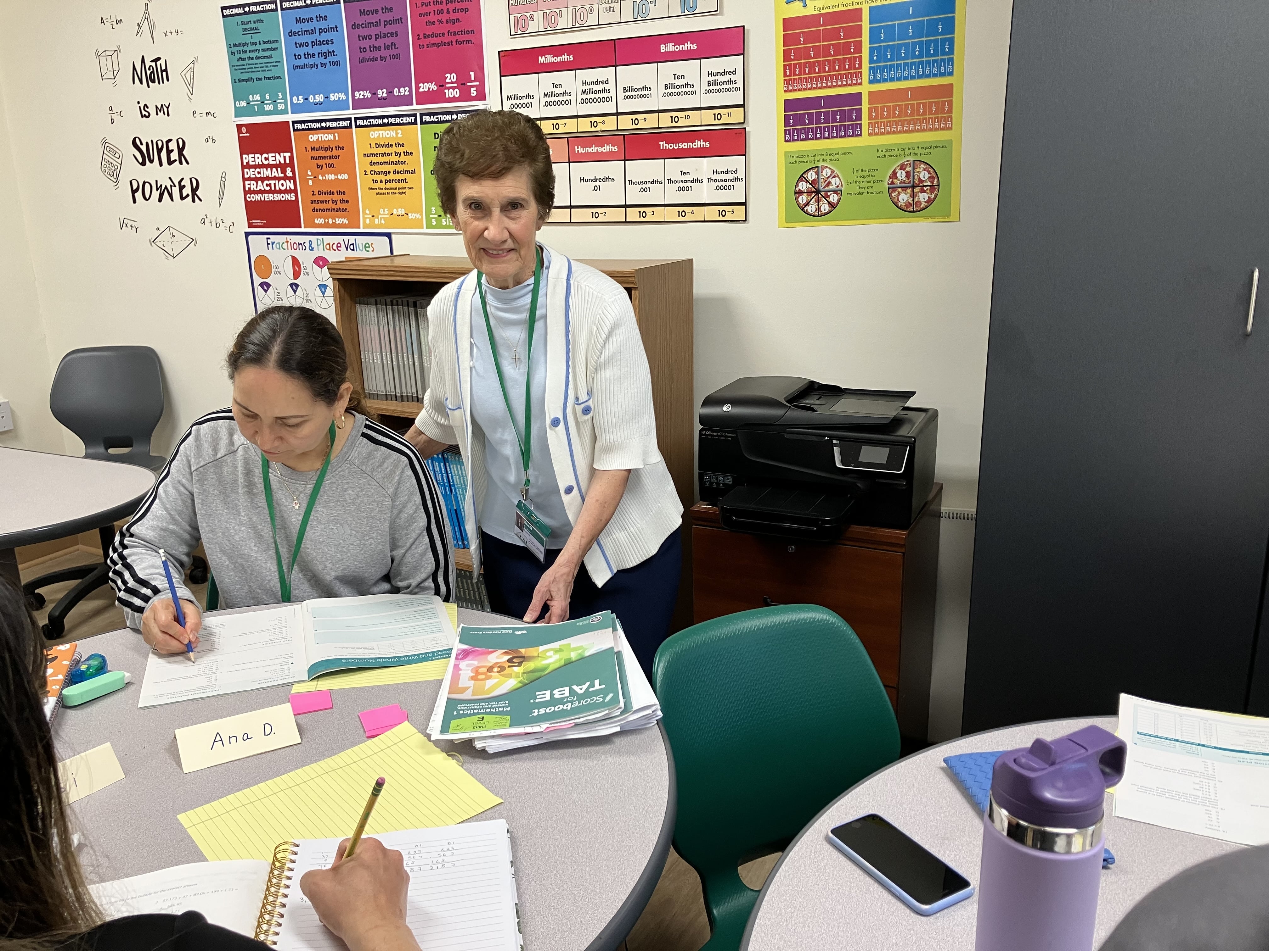 Standing beside the table, Sister Jeanne Amore watches as a student sits and writes on a sheet of paper.