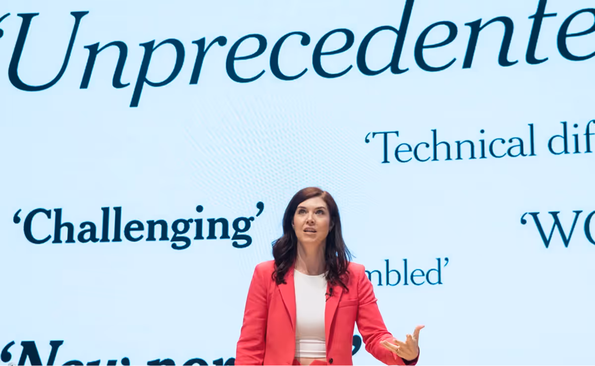 A woman in a red jacket stands in front of a screen with words like "imprecedented" and "technical" on it.