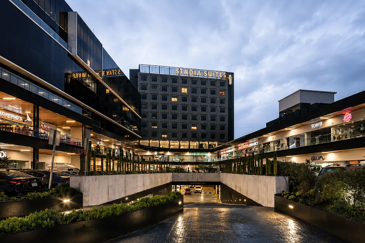 Plaza Ubika en Universidad, Querétaro, al atardecer: hotel Stadia Suites, tiendas iluminadas y entrada al estacionamiento subterráneo.