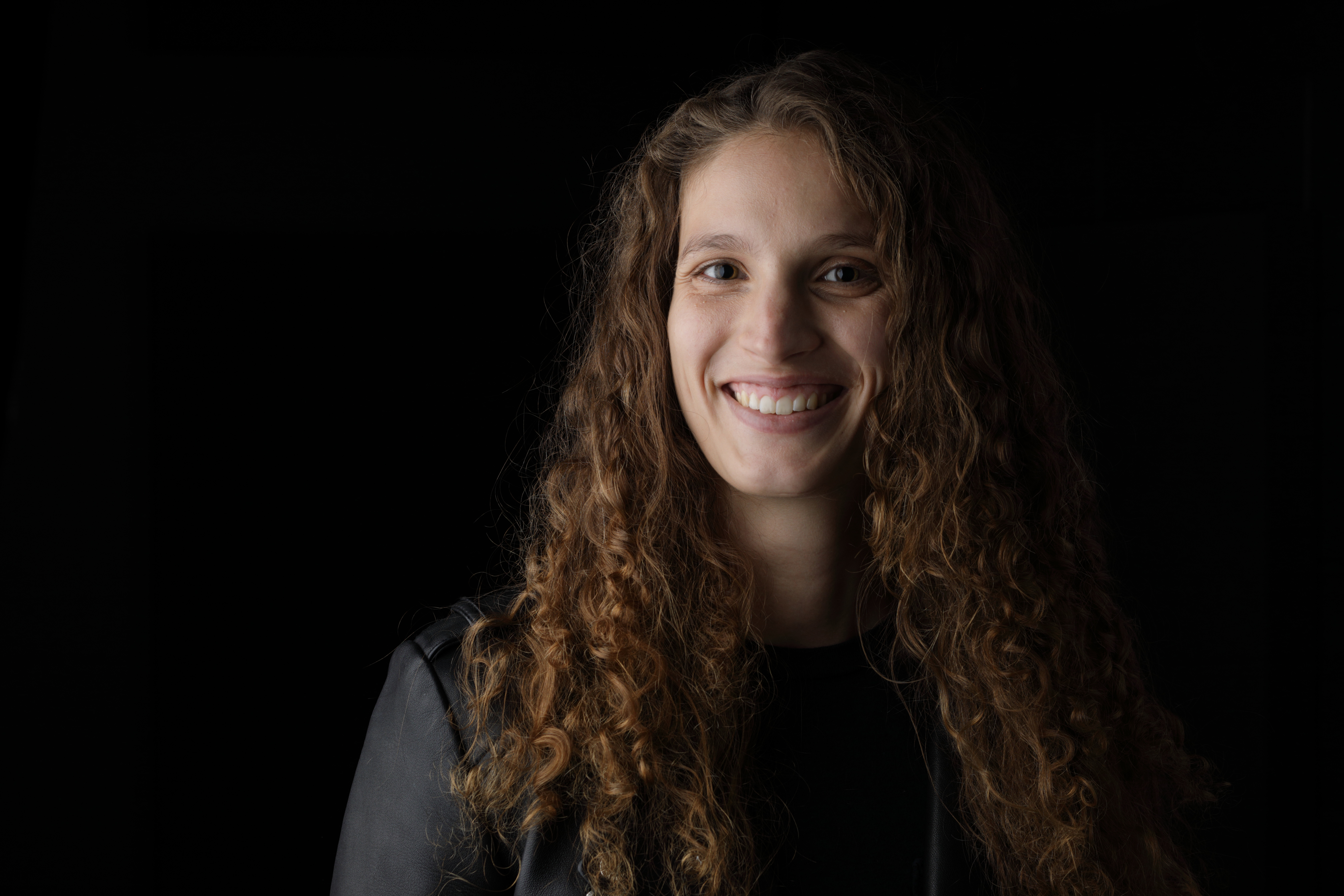 Natasha, a white woman with curly brown hair smiles at the camera. She is wearing a black leather jacket and the backdrop of the photo studio is dark.