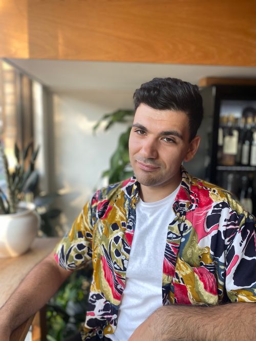 Arman, a brown journalist with stylish black hair, sits in a room in a colourful overshirt with a white one under it.
