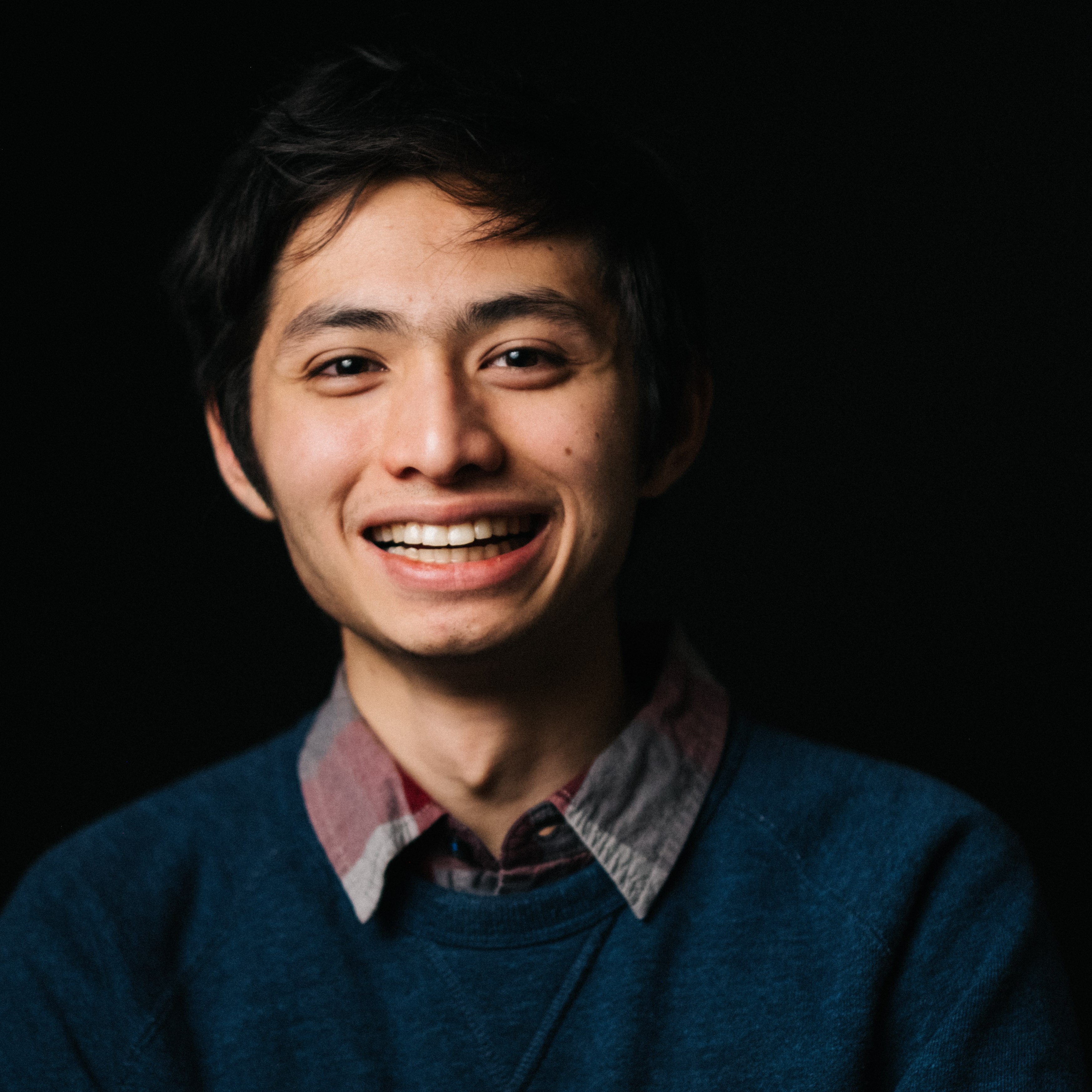 Aloysios, a Filipino man, smiles at the camera from in front of a black background He is wearing a blue sweater and a purplish coloured shirt