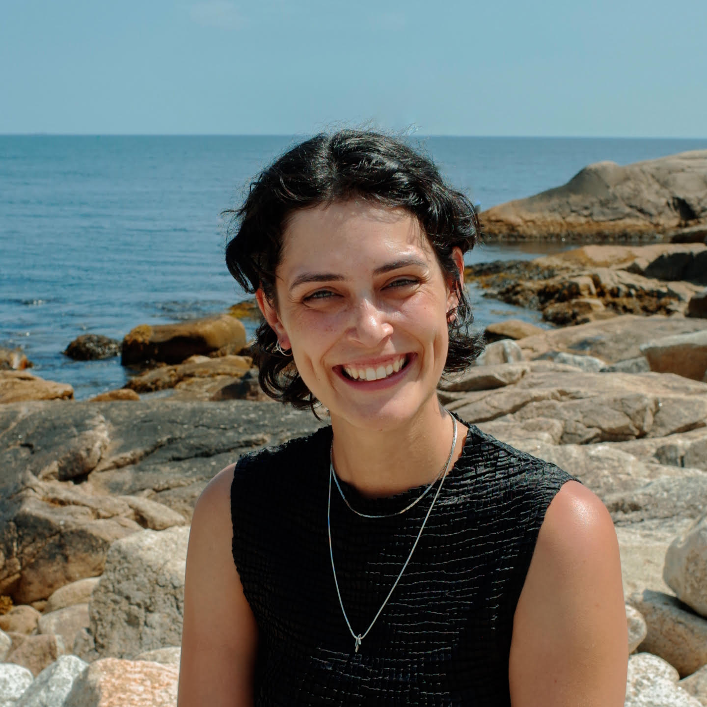 A photograph of Cloe, smiling with rocks and ocean behind her. She is wearing a black shirt and silver necklaces.