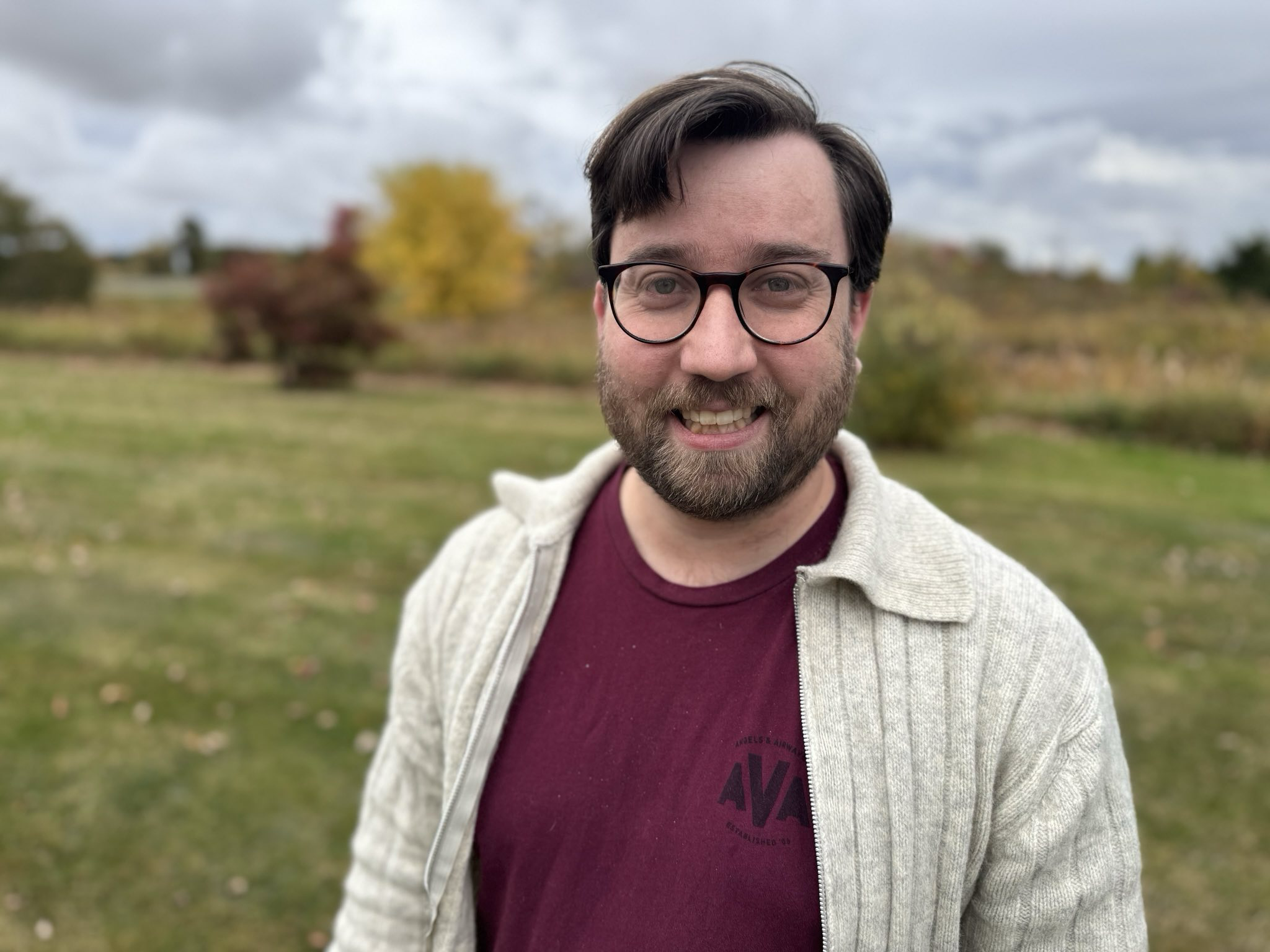 Lambert, a white media librarian stands in front of a park in a red shirt and white sweater. He is smiling with glasses. The field behind him is in fall colours