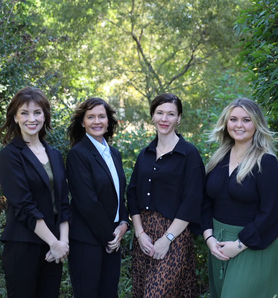 Four women standing outdoors in front of green trees, smiling and dressed in business casual attire.