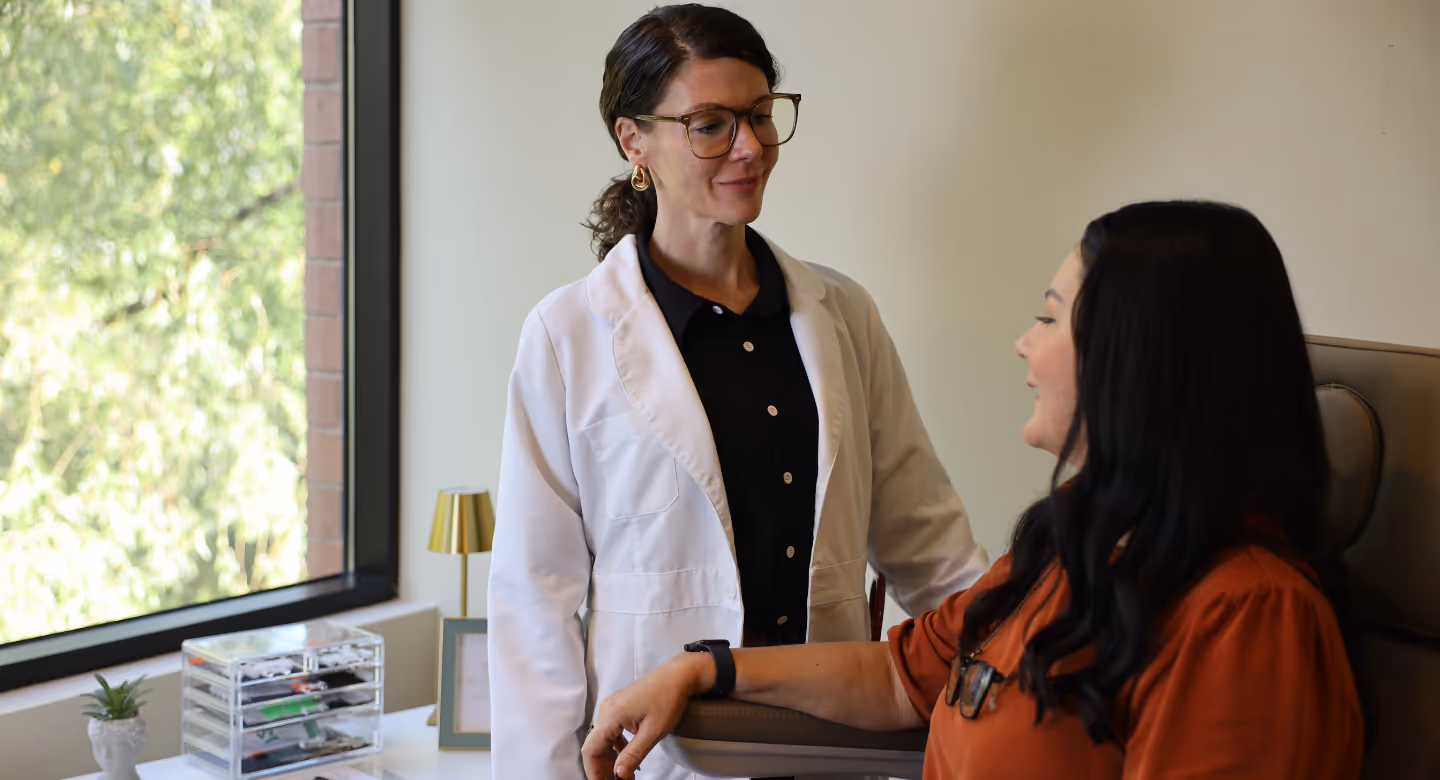 Female doctor in a white coat talking with a seated woman in an orange top in a bright office.