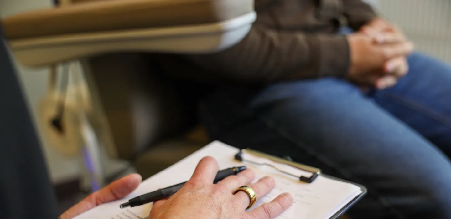Close-up of a hand holding a pen over a clipboard with papers, a person with clasped hands wearing blue jeans sits in the background.