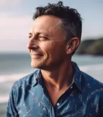 Middle-aged man with short curly hair smiling and looking to the side near a beach at sunset.