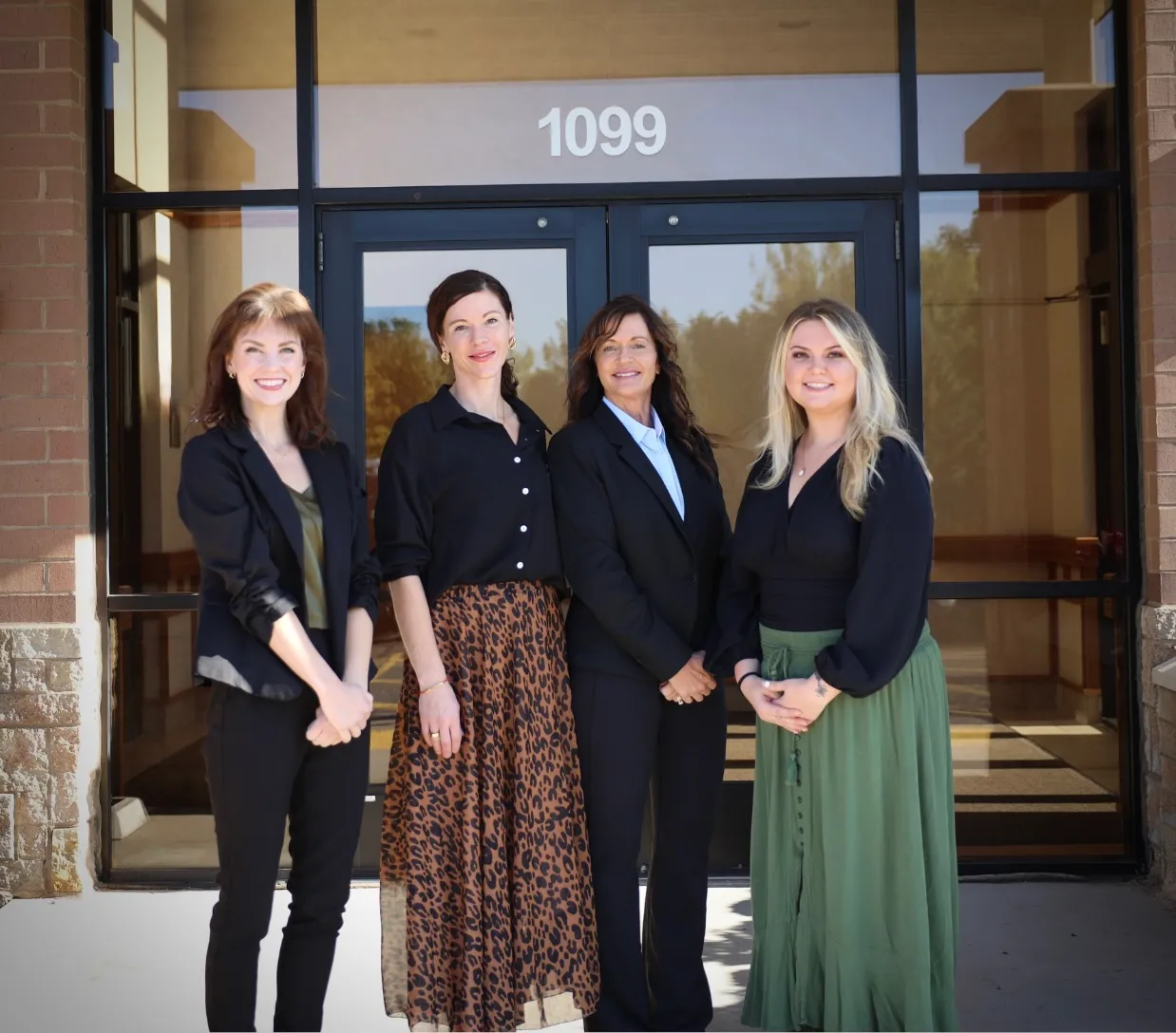 Four women standing outside building entrance with glass doors labeled 1099, smiling and dressed in business casual attire.