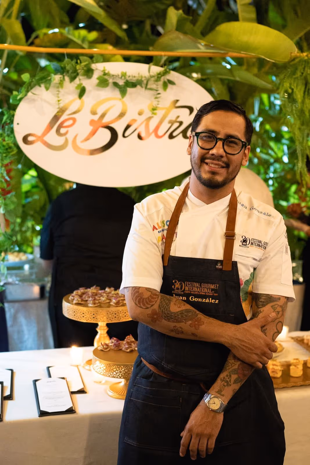 Smiling chef with glasses and tattoos wearing a Festival Gourmet International apron in front of a table with desserts and a 'Le Bistro' sign.