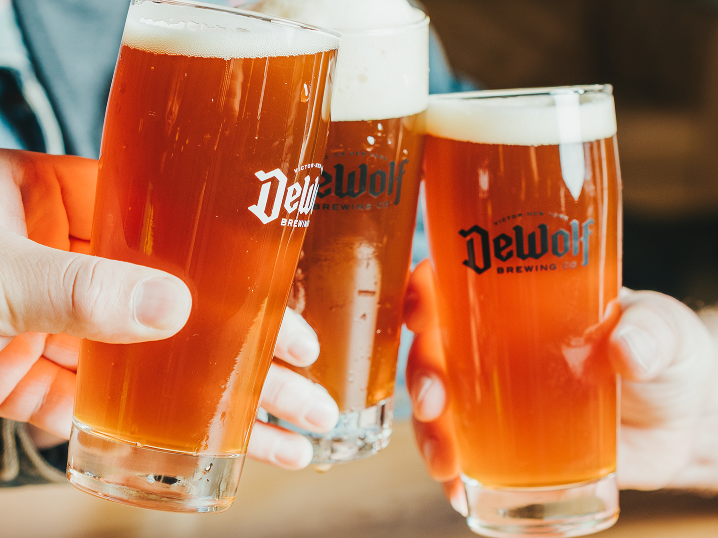 Three pints of Volksbier Vienna lager from DeWolf Brewing Company, amber-colored with a creamy white head, clinking together.