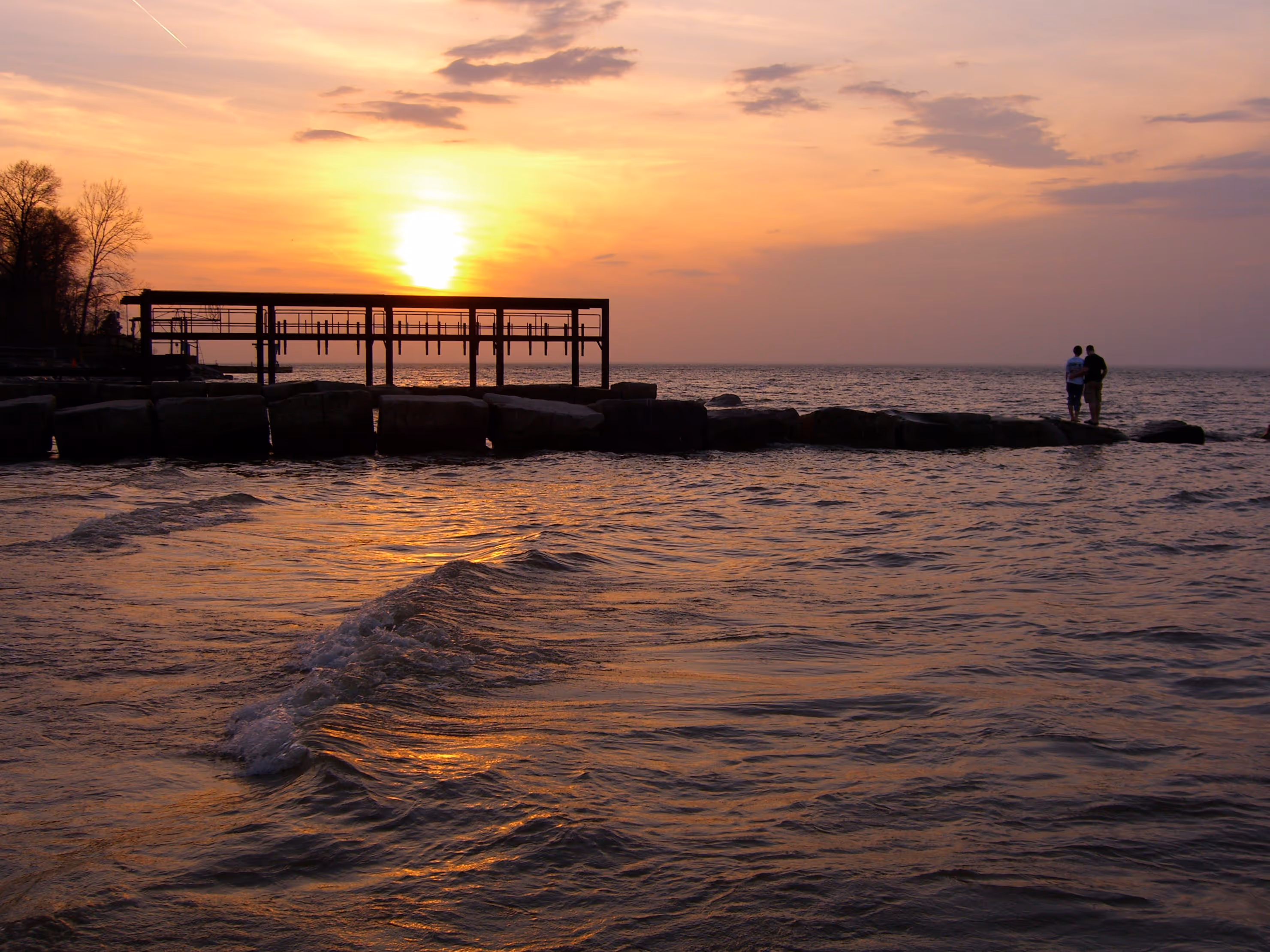 Sunset over water with a wooden pier and two people standing on rocks by the shore.
