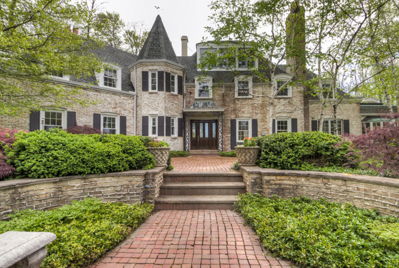 Large brick house with a central turret, multiple windows with black shutters, and a red brick pathway leading to double front doors.