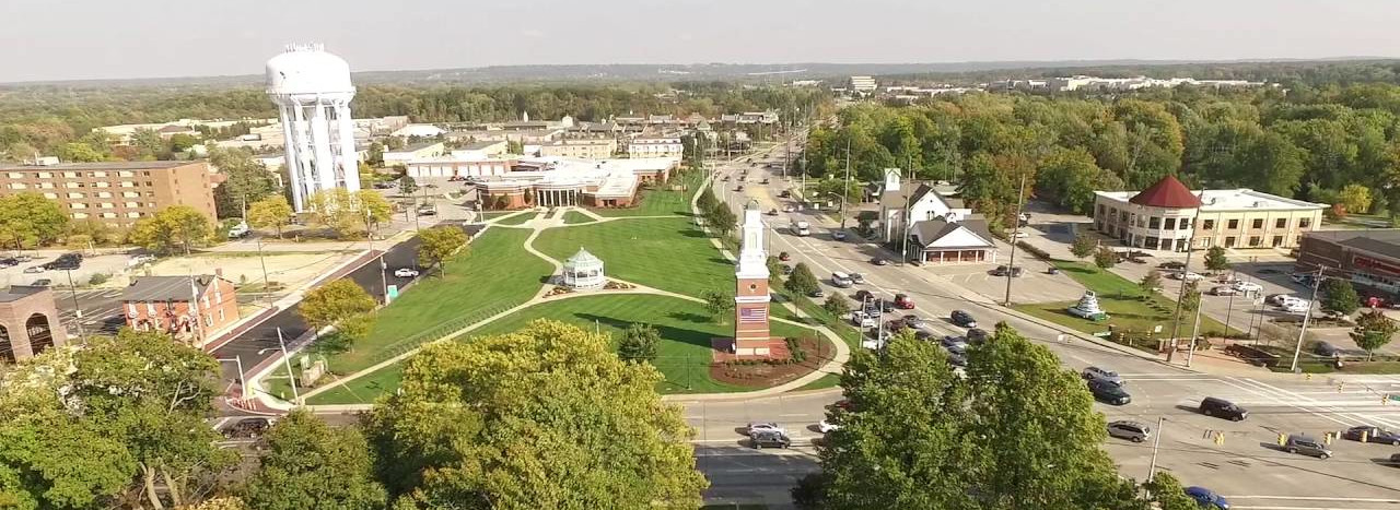 Aerial view of a suburban area with a green park featuring a gazebo and clock tower, surrounded by roads, trees, and buildings under a clear sky.