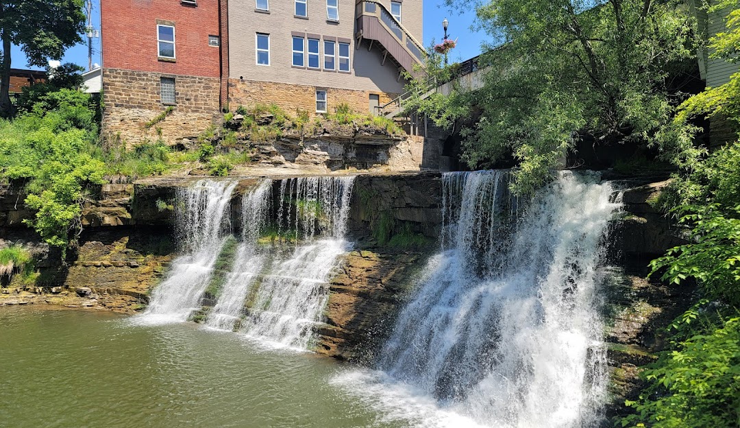 Waterfalls flowing over rock ledges into a river, with trees and buildings in the background under a clear blue sky.