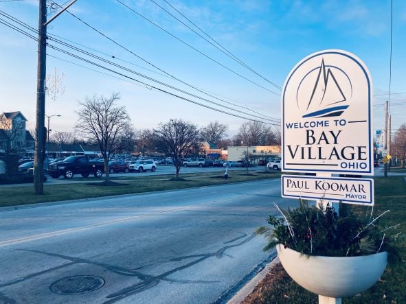 Street view of Bay Village, Ohio welcome sign with a planter, trees, and parked cars under a clear blue sky.