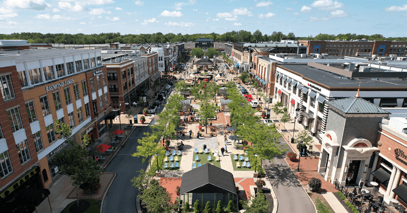 Aerial view of a bustling urban outdoor shopping and dining area with tree-lined pedestrian walkways, benches, and people enjoying the space.