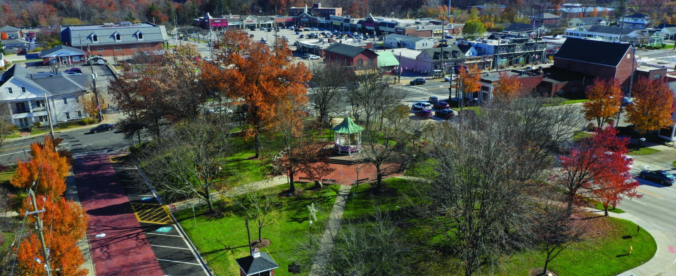 Aerial view of a small town park with a gazebo surrounded by trees with autumn foliage and nearby streets with cars and buildings.