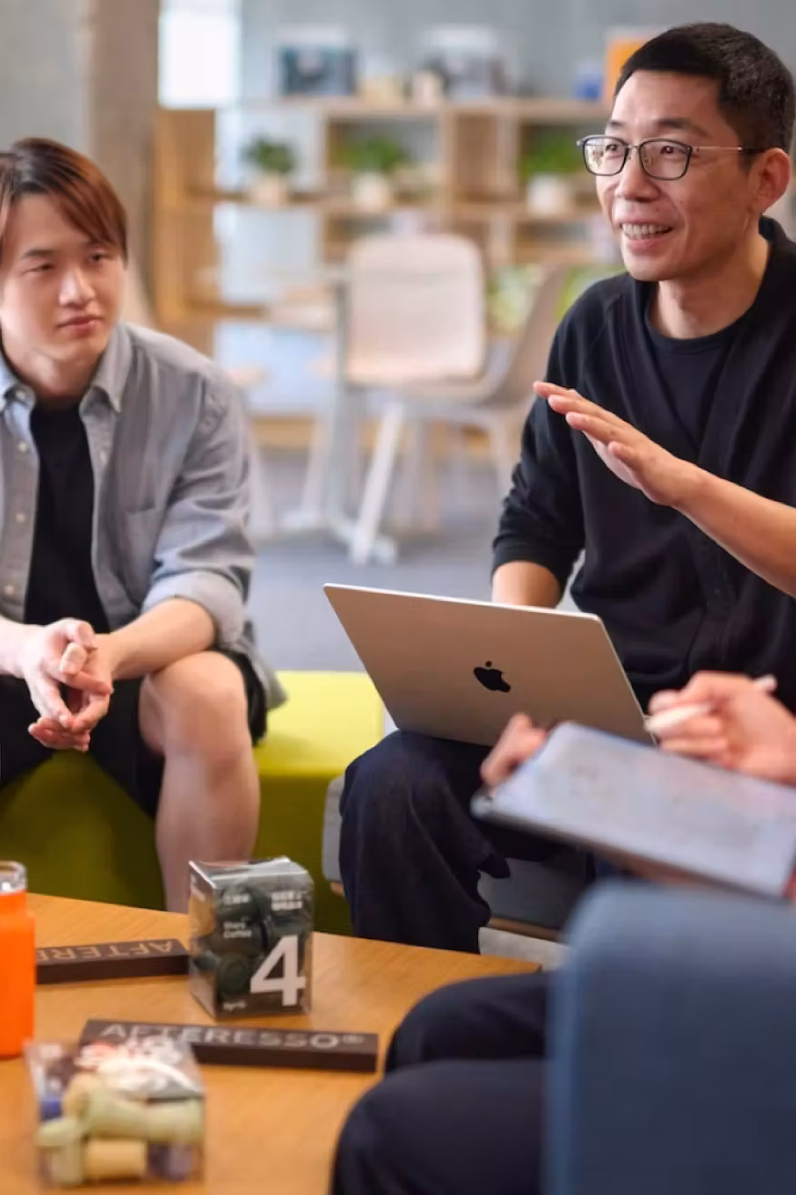 กลุ่มคนหนุ่มสาวนั่งคุยกันในห้องทำงานแบบเปิด พร้อมแล็ปท็อปและบันทึกในมือ