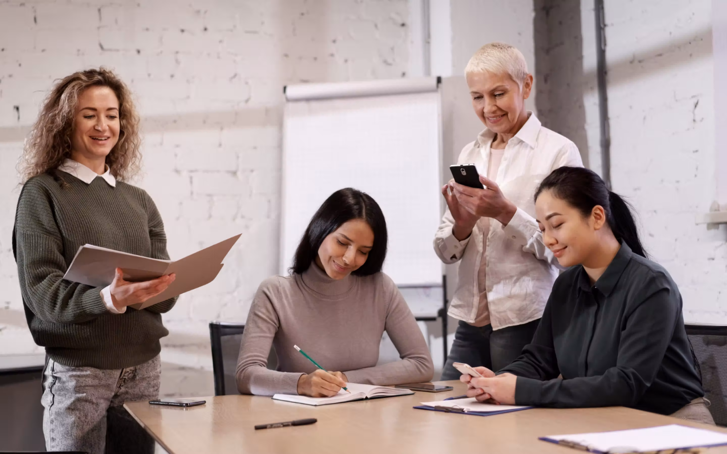 ผู้หญิงสี่คนทำงานร่วมกันในห้องประชุม โดยสองคนใช้โทรศัพท์มือถือ หนึ่งคนเขียนบันทึก และอีกคนยืนอ่านแฟ้มเอกสาร