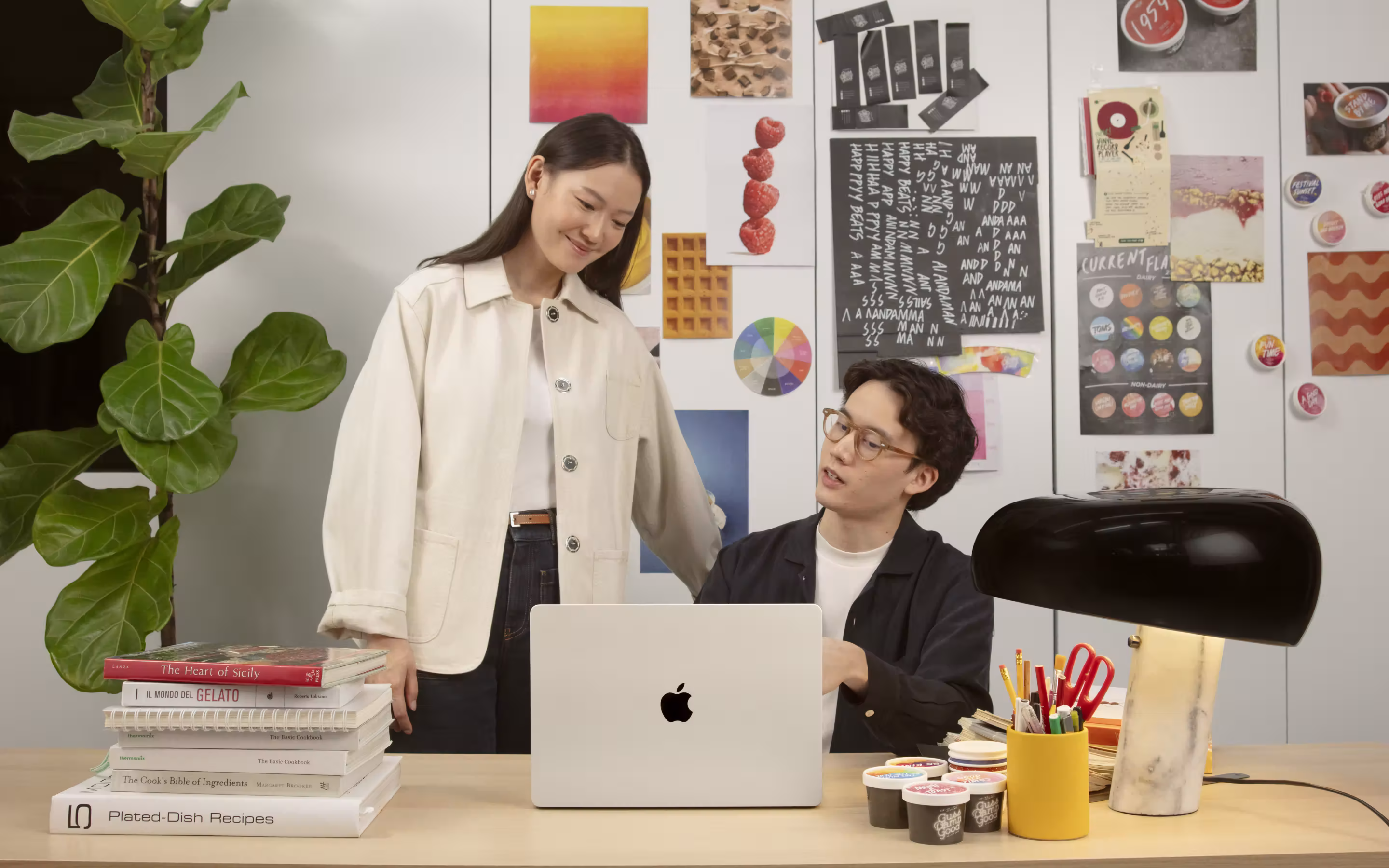 ผู้หญิงยืนยิ้มมองผู้ชายที่นั่งทำงานที่โต๊ะพร้อมแล็ปท็อป Apple และของใช้บนโต๊ะในห้องที่มีภาพและโน้ตติดบนผนัง