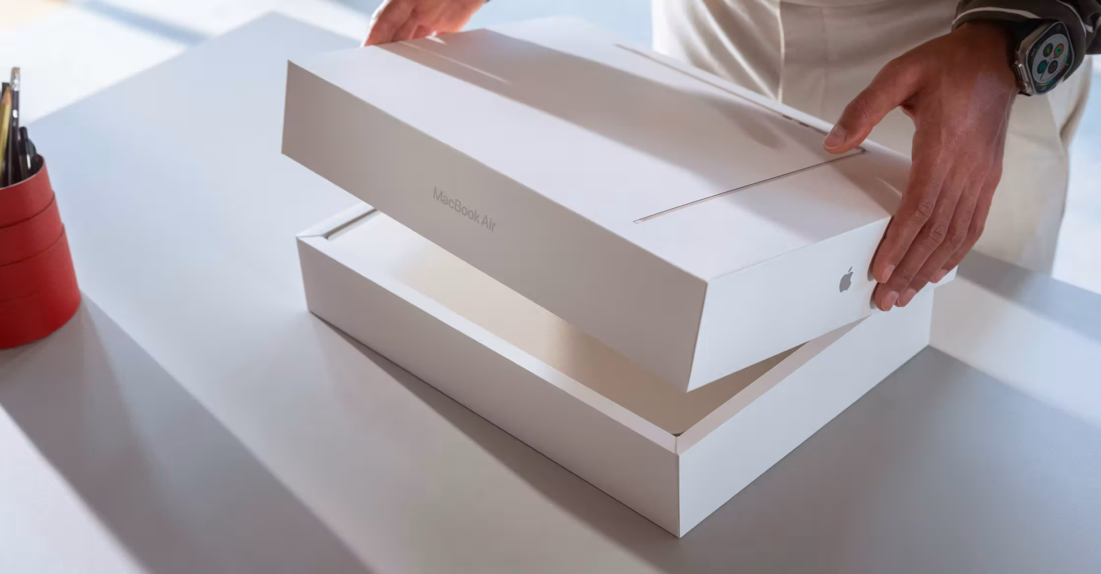 มือกำลังเปิดกล่อง MacBook Air สีขาวบนโต๊ะพร้อมแก้วใส่ปากกาอยู่ข้างๆ
