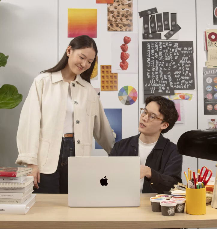 ชายหญิงสองคนทำงานร่วมกันที่โต๊ะโดยมีแล็ปท็อป Apple และของใช้ในสำนักงานหลากหลายรายการ