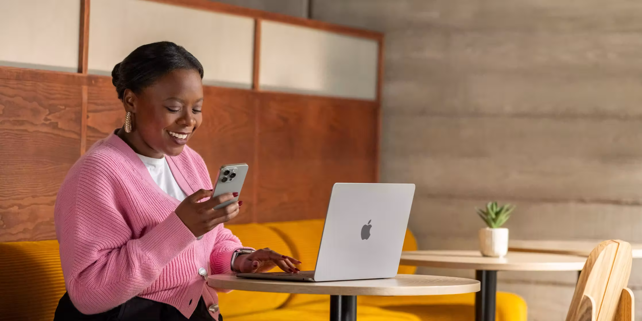 หญิงสาวสวมเสื้อคาร์ดิแกนสีชมพูกำลังยิ้มขณะใช้โทรศัพท์มือถือและแล็ปท็อปในร้านกาแฟ