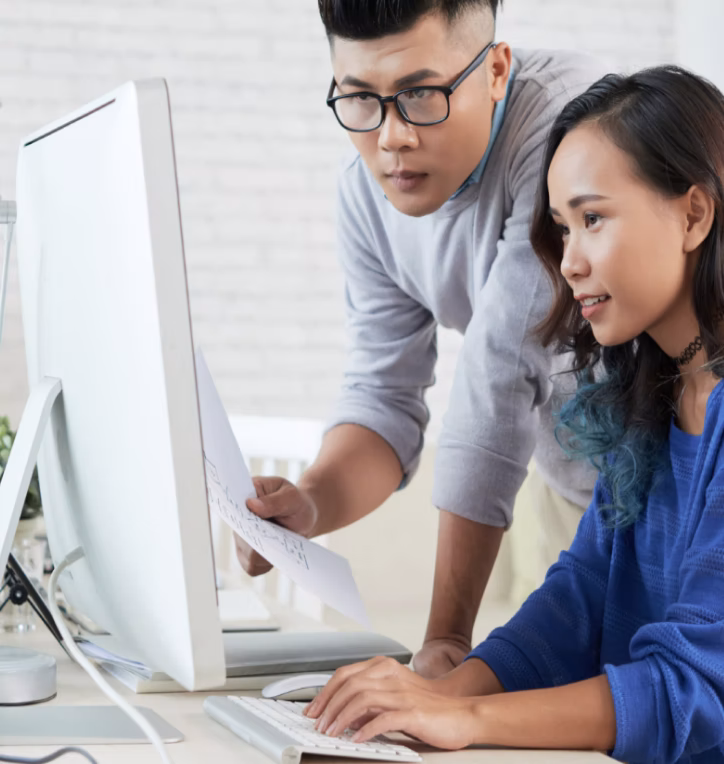 ชายและหญิงชาวเอเชียกำลังทำงานร่วมกันโดยดูที่หน้าจอคอมพิวเตอร์และผู้ชายถือกระดาษในมือ