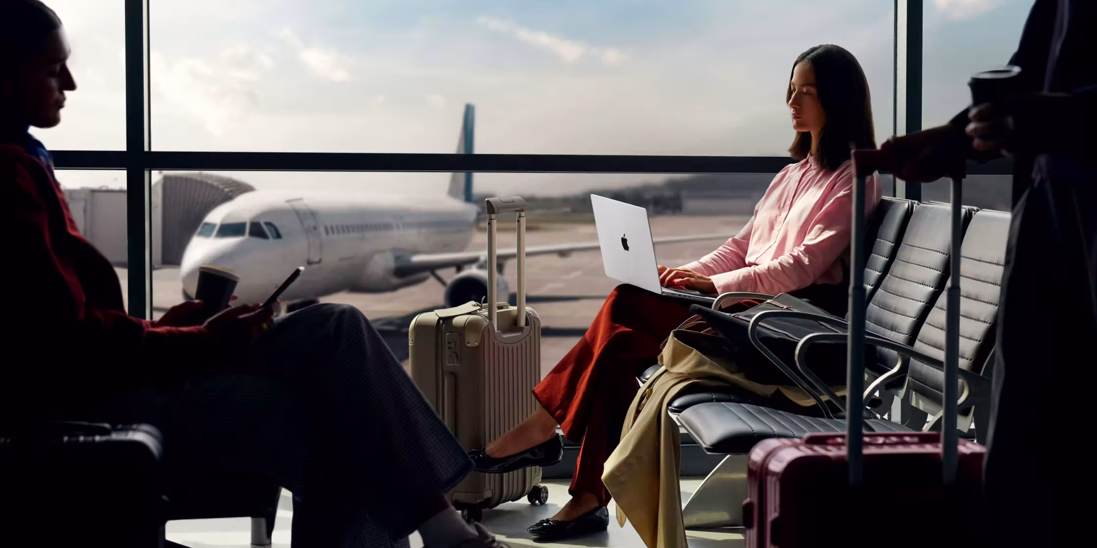 ผู้หญิงนั่งรอที่สนามบินใช้แล็ปท็อปและผู้โดยสารคนอื่นถือกระเป๋าลากใกล้เครื่องบินจอดอยู่ด้านนอก