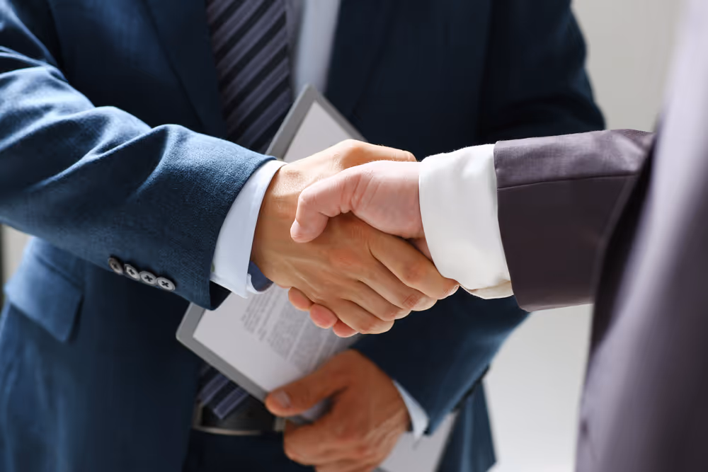 Two businessmen shaking hands, one holding a clipboard with documents.