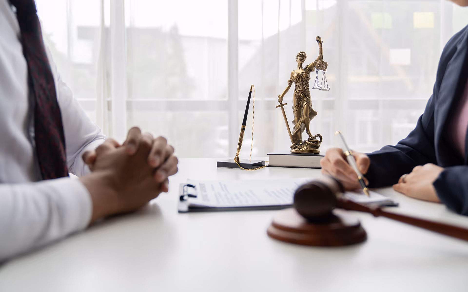 Two people at a desk in a legal consultation setting with a Lady Justice statue, a pen on a stand, a clipboard with documents, and a judge's gavel.
