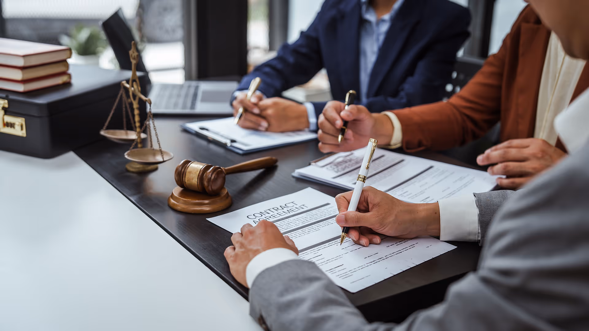 Three people signing contract agreement documents at a desk with a judge's gavel and scales of justice.