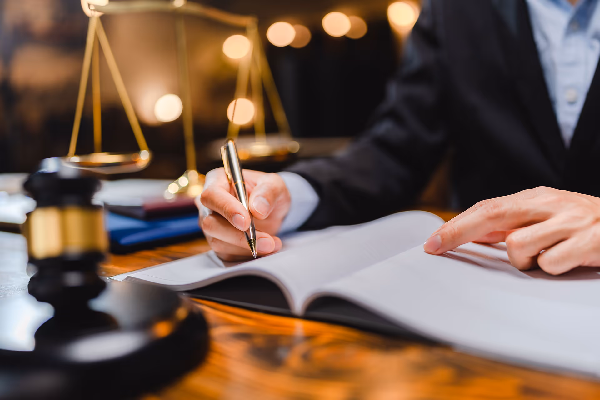 Person in suit writing in an open book with legal scales and a gavel blurred in the background.