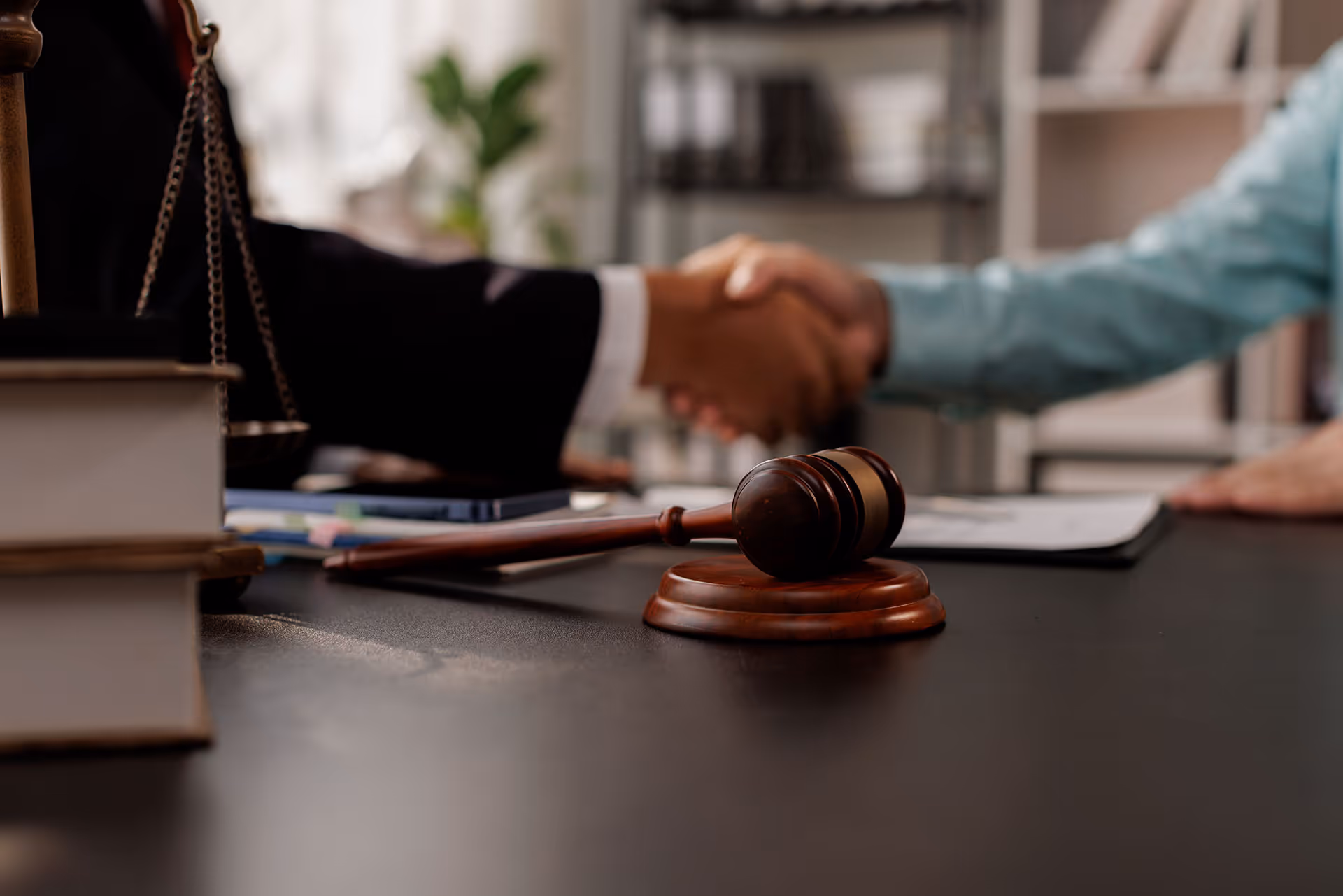 Close-up of a wooden judge's gavel on a desk with two people shaking hands in the background.